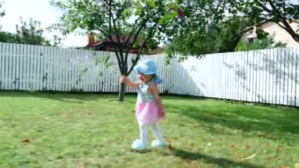 a pretty one-year-old girl in a hat from the sun and a gorgeous dress, goes quietly to her mother. Mom takes baby in her arms, turns around. In the summer, in the garden. - Video