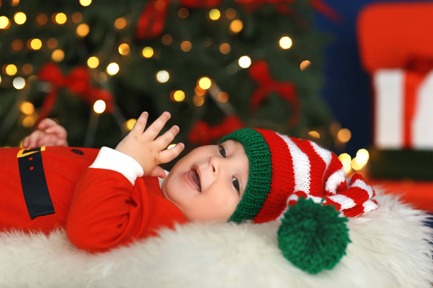Cute little baby in Santa costume lying against blurred Christmas lights background - Foto, imagen