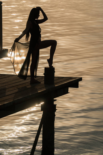 Tyttö uimapuvussa ja viitta päällä seisoo laiturilla ja kohtaa aamunkoiton.
 - Valokuva, kuva