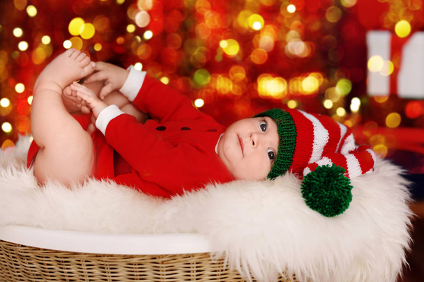 Cute little baby in Santa costume lying against blurred Christmas lights background - Fotografie, Obrázek