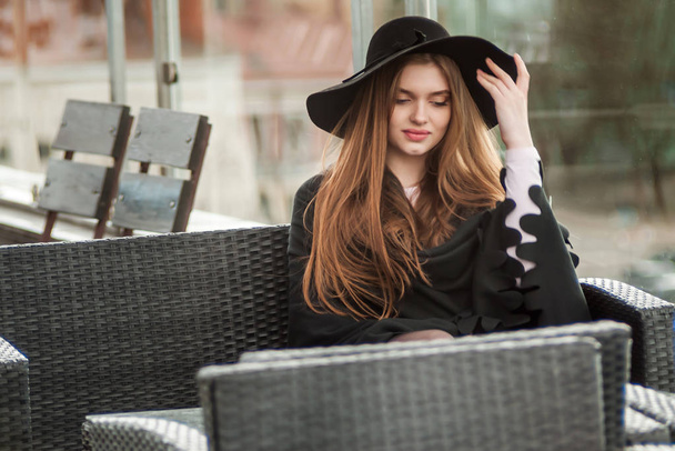 girl in a black wide-brimmed hat and cloak sitting on the terrace of the restaurant - Photo, image
