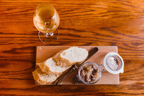 The horizontal above view of the food composition consisted of the bread and jar of meat on the wooden board standing near the glass with white wine. - Foto, imagen