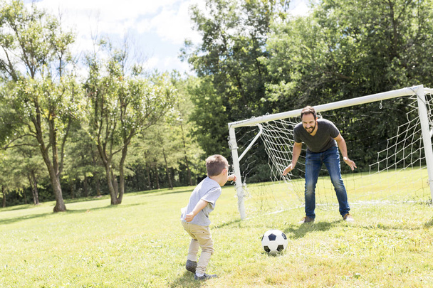 Ο νεαρός πατέρας με τον μικρό του γιο παίζουν ποδόσφαιρο στο γήπεδο ποδοσφαίρου. - Φωτογραφία, εικόνα