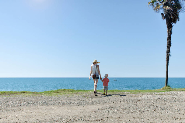 Anya és fia áll háttal a magas pálma fa, a tenger és a kék ég háttér - Fotó, kép