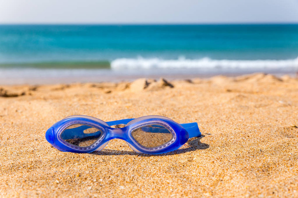 Gafas de natación azules tumbadas en la playa con mar
 - Foto, Imagen