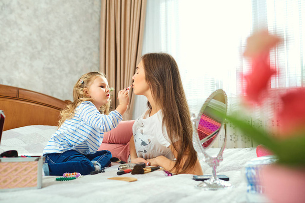 Mother and daughter do make-up, play on the bed by the window. - Zdjęcie, obraz