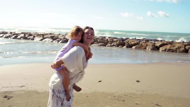 Mujer sosteniendo a su hijita en la espalda sonriendo frente al océano feliz - Metraje, vídeo
