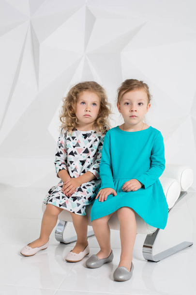 zwei kleine Freundinnen in identischen Kleidern in verschiedenen Farben sitzen auf einem Stuhl in einem Studio mit weißen Wänden - Foto, Bild
