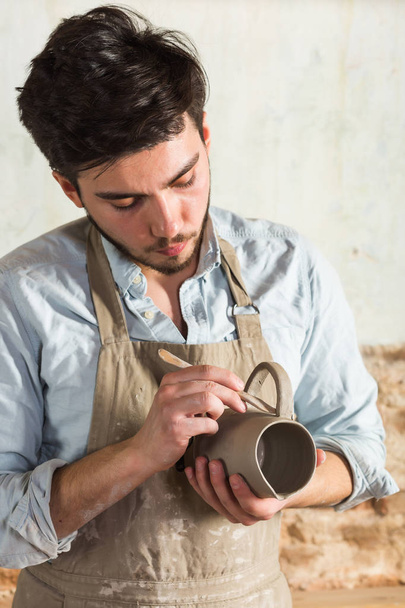 potter, workshop, ceramics art concept - young craftsmans hands with unbaked clay cup, male master standing at a workplace, ceramist hold the unfinished jug and tools, vertical. - Photo, image