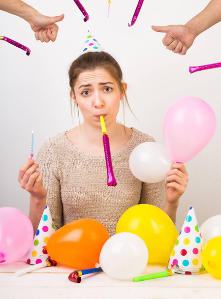 emotions, party, birthday. young beautiful girl dressed in beige cardigan and colorful party hat, she is surrounded by balloons and whistles, but looks really sad - Foto, Imagem