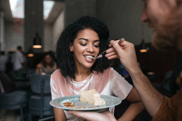 Young African American girl with dark curly hair eating cake in restaurant. Portrait of young man feeding his girlfriend at cafe - Photo, Image