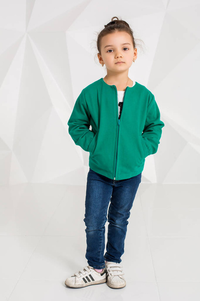 Beautiful little girl in a green jacket and jeans posing in front of camera in studio - Photo, Image