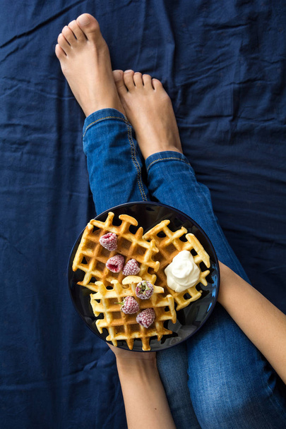Belgian waffles with cream and frozen raspberries on blue ceramic plate in woman' s hands. Top view - 写真・画像