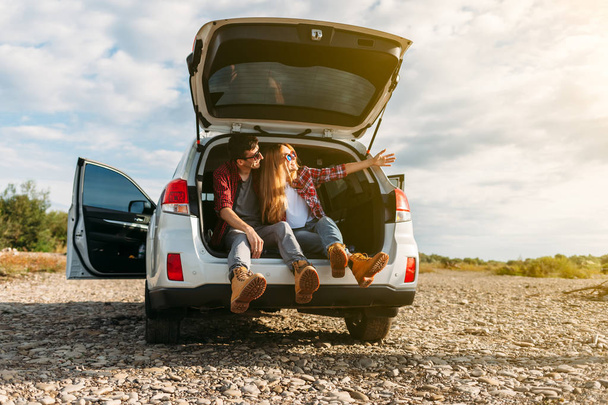 Happy traveler couple sitting in car open trunk and watch the sunrise - Photo, Image