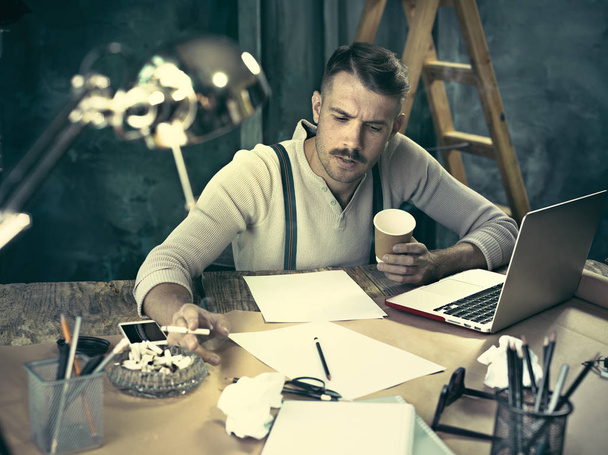 Красивий елегантний чоловік сидить за домашнім столом, працює і використовує ноутбук під час куріння сигарет
 - Фото, зображення