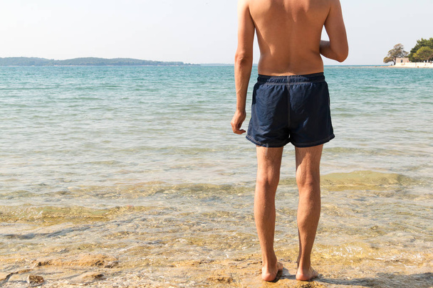 Masculine model of backs looking at the sea. Clear water, bright sun and sunscreen for sunburn. Man in blue shorts getting ready to go swimming in the mediterranean sea. - Photo, Image
