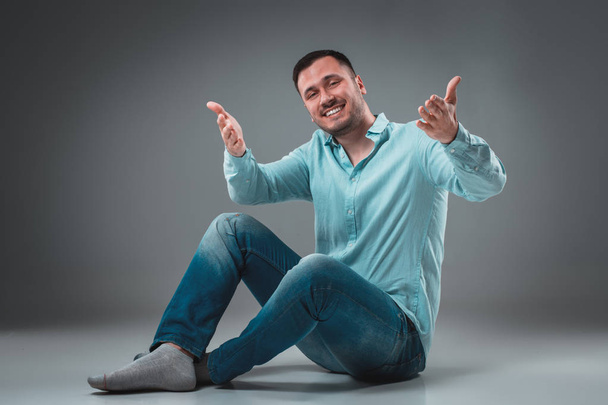 schöner junger Mann sitzt mit erhobenen Händen auf dem Boden und gestikuliert glücklich auf grauem Hintergrund - Foto, Bild