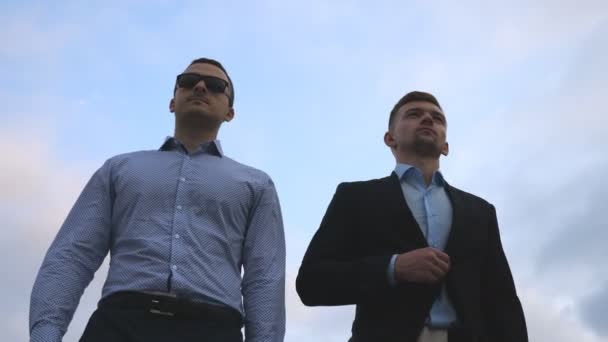 Két fiatal üzletemberek séta a városban a kék ég háttér. Üzletemberek commuting-hoz dolgozik együtt. Magabiztos srácok iroda útját is. Kollégák lesz szabadtéri. Lassú mozgás közelről - Felvétel, videó