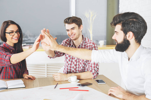 Young team members hi-fiving each other at modern workplace with blurry city view. Coworking concept  - Фото, изображение