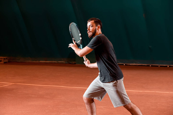 Pohledný mladý muž v tričko hospodářství tenisové rakety na tenisový kurt - Fotografie, Obrázek