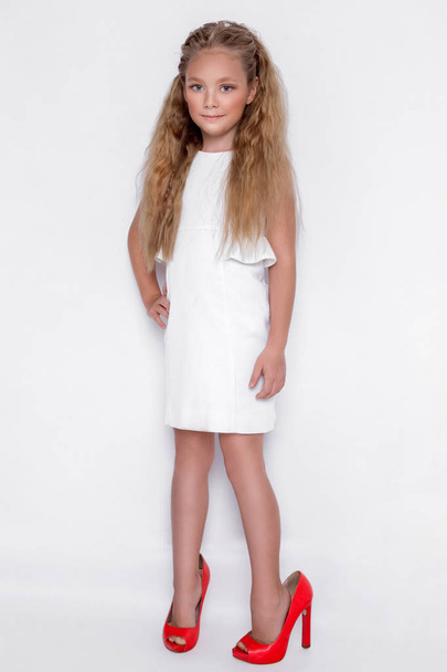 Досить приголомшлива маленька дівчинка принцеса, що стоїть на білому тлі в білій сукні, і вона має велике взуття на ногах
 - Фото, зображення