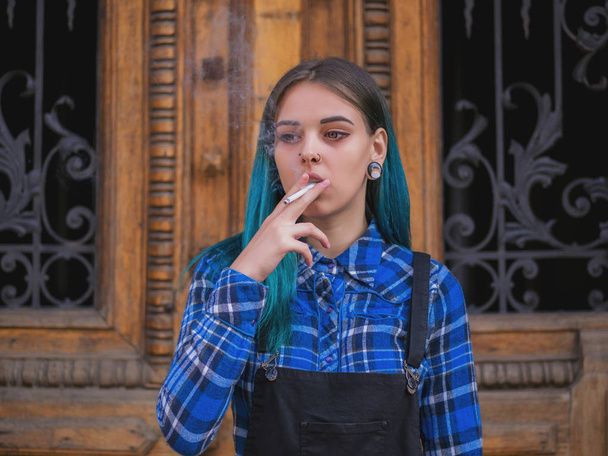 Young punk girl smoking cigarette by the wooden door.Hipster with blue dyed hair. Woman with piercing in nose, violet lenses, ears tunnels and unusual hairstyle. - Foto, afbeelding