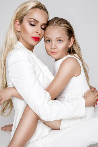 Stunning beauty a young mother with a cute blonde daughter standing on a white background dressed in a  spring clothing - Photo, Image