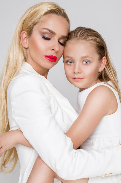 Stunning beauty a young mother with a cute blonde daughter standing on a white background dressed in a  spring clothing - Φωτογραφία, εικόνα