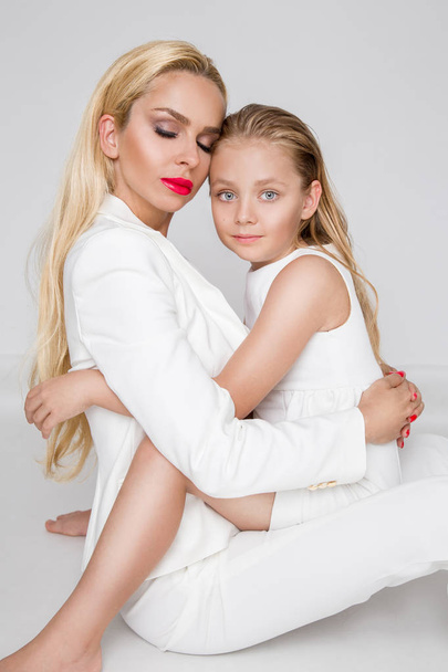 Impresionante belleza una madre joven con una linda hija rubia de pie sobre un fondo blanco vestido con una ropa de primavera
 - Foto, imagen