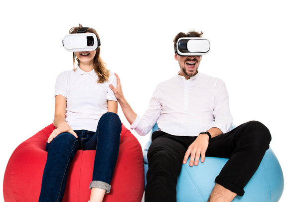 Excited young couple experiencing virtual reality seated on beanbags isolated on white background - 写真・画像