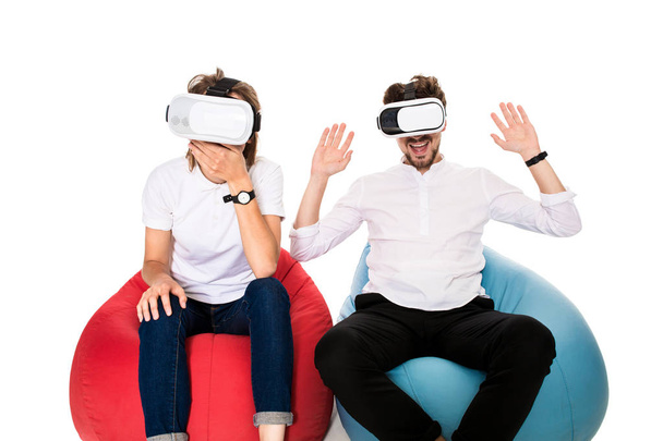 aufgeregtes junges Paar erlebt Virtual Reality sitzend auf Sitzsäcken isoliert auf weißem Hintergrund - Foto, Bild