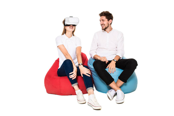 Uśmiechający się przyjaciółmi przeżywa siedzi na beanbags izolowana na białym tle okulary wirtualnej rzeczywistości. - Zdjęcie, obraz