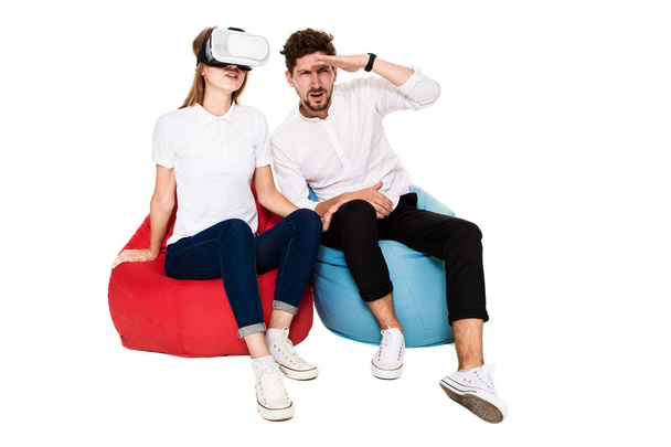aufgeregtes junges Paar erlebt Virtual Reality sitzend auf Sitzsäcken isoliert auf weißem Hintergrund - Foto, Bild