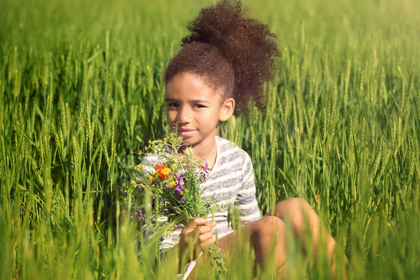 Little Afro American girl with bouquet of wildflowers in green field - Фото, изображение