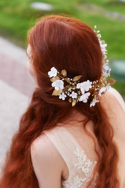 Beautiful red-haired bride sits back .Slightly visible profile. Long curly hair with handmade jewelry. - Fotó, kép