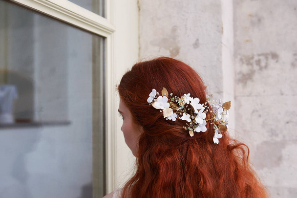 Beautiful red-haired bride sits back .Slightly visible profile. Long curly hair with handmade jewelry. - Photo, Image