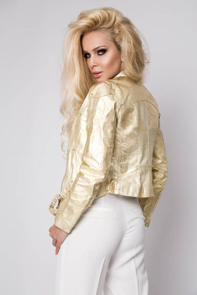 Красивая сексуальная блондинка с длинными волосами, стоящая на белом фоне в золотой кожаной куртке байкера стиле на велосипеде и чувственно, заманчиво смотрит в объектив
 - Фото, изображение
