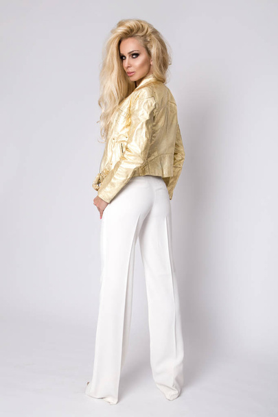 Красивая сексуальная блондинка с длинными волосами, стоящая на белом фоне в золотой кожаной куртке байкера стиле на велосипеде и чувственно, заманчиво смотрит в объектив
 - Фото, изображение