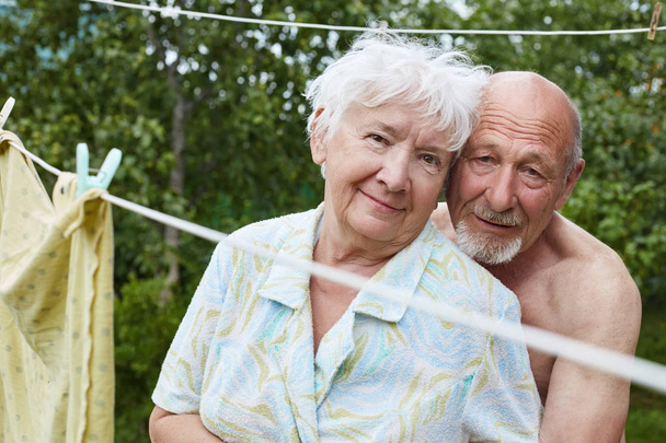 authentische Aufnahme eines alternden Ehepaares im russischen Dorfhof. Der braungebrannte Mann mit dem grauen Bart umarmt seine Partnerin mit Liebe und Hingabe, die Frau lächelt mit Zärtlichkeit.. - Foto, Bild