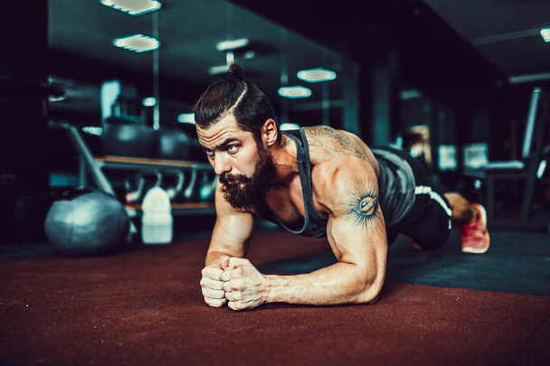 Muscled young man wearing sport wear and doing plank position while exercising on the floor in loft interior - Foto, Imagen