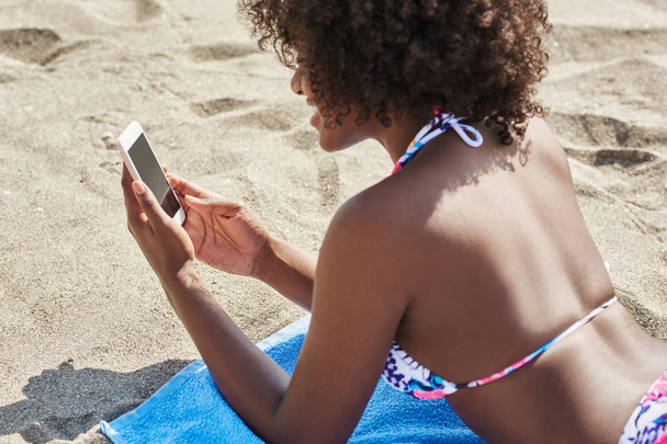 Afro american woman lying on beach using smartphone - Foto, Imagen