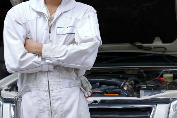 Ritratto di giovane meccanico fiducioso in uniforme con braccia incrociate e in piedi contro l'auto in cappa aperta al garage di riparazione
. - Foto, immagini