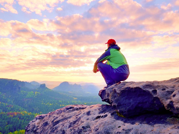 Vista traseira do turista adulto em casaco preto verde sentado no pico rochoso enquanto desfruta de vista maravilhosa f
 - Foto, Imagem