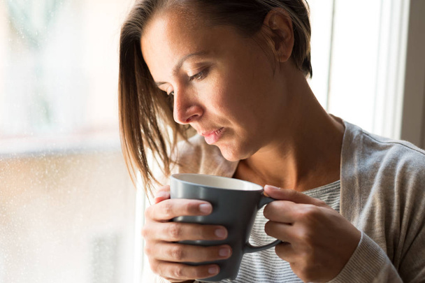 Woman holding a cup of coffee portrait next to a window  - Zdjęcie, obraz