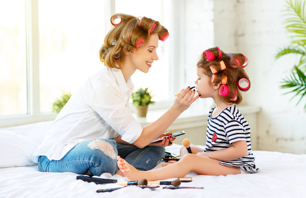 Mother and baby daughter in curlers  doing makeup on bed - Photo, Image
