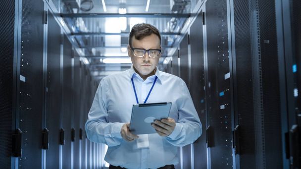 IT Technician Walks Through Rows of Server Racks in Data Center. Simultaneously He Works on a Tablet Computer. - 写真・画像