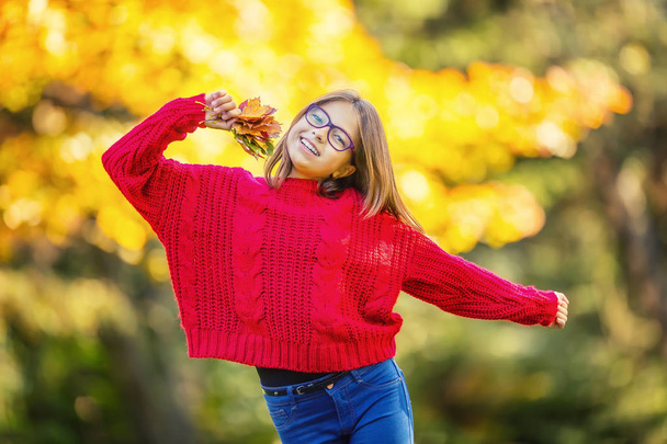 Happy fall girl smiling and joyful holding autumn leaves. Beautiful young girl with maple leaves in red cardigan - 写真・画像