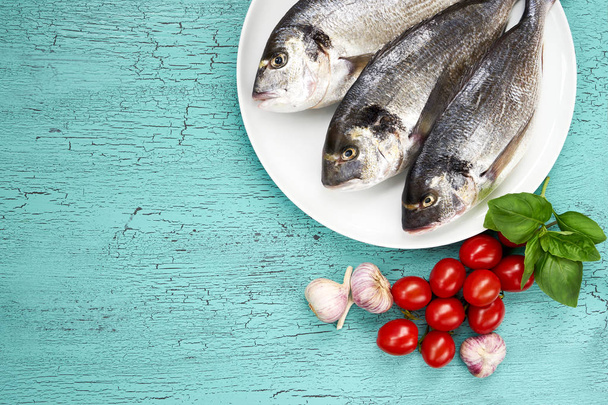 roher frischer Dorado-Fisch auf weißem Teller und Gemüse auf blauem Tisch. Ansicht von oben - Foto, Bild