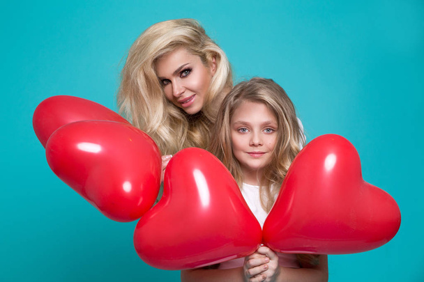 schöne blonde Mutter und Tochter stehen auf blauem Hintergrund und halten rote Valentinusballons in der Hand - Foto, Bild