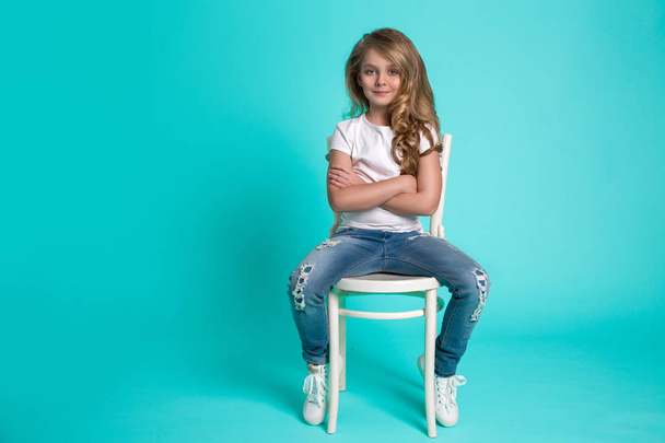Beautiful stunning little blonde girl in denim jeans sitting on a chair on a blue background - Фото, изображение